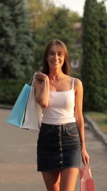 Elektrikli mavi kot etek giyen bir kadın, alışveriş torbalarıyla sokakta yürürken gülümseyerek ve kollarıyla el kol hareketi yaparak.