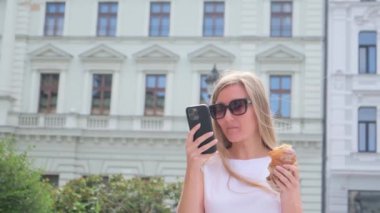 Uzun saçlı güzel bir genç kız donut yiyor ve güzel bir binanın arka planında telefona bakıyor. Güzel mimarinin arka planındaki kız..
