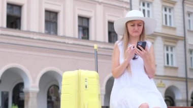 Güzel sarışın bir turist şehirdeki bir binanın arka planında oturuyor ve telefonda bir şey arıyor. Beyaz elbiseli genç turist bayan.