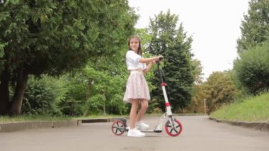 Elektrikli scooter kullanan küçük bir kız. Tekerlekleri asfaltta dönmüyor. Bulutlu bir gökyüzünün altında yol boyunca ağaçlar ve bitkiler.