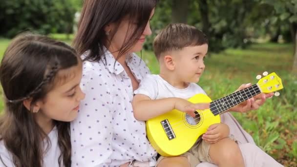 一个女人和两个孩子一起坐在草地上 分享着音乐的欢乐 玩的很开心 一把小吉他会给孩子们带来乐趣和闲暇 — 图库视频影像