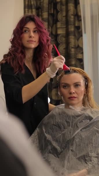 女性は サロンで美容師によって髪を染めている長いブロンドの髪を得ています 美容師は慎重に染料を適用していますが 女性は鏡の新しい髪型をジェスチャーし 認めています — ストック動画