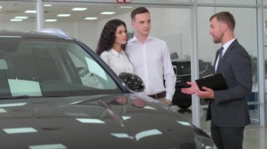 Bir erkek ve bir kadın galeride lüks bir arabayı inceliyor, hayranlık jestlerini paylaşırken dış, kaput ve otomotiv aydınlatma araçlarını inceliyor.