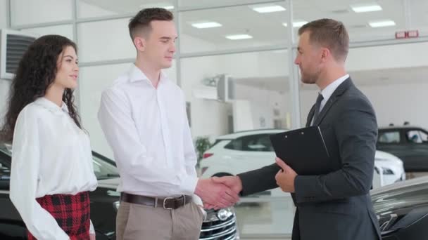 ドレスシャツを着た男性が ショールームにいる女性と男性と車両について話し合っている 彼らは車のディーラーで新しい車を購入しながら微笑んでいる — ストック動画