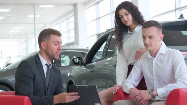 フォーマルウェアのカップルは 車のショールームでラップトップを見ています 彼らが購入に興味を持っている車の自動車のデザインと機能を議論 — ストック動画
