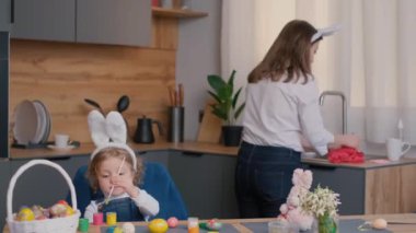 Küçük bir kız masada paskalya yumurtalarını süslerken bir kadın mutfakta bulaşık yıkıyor. Evin iç tasarımı çiçek süslemelerini içerir.