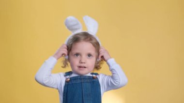 Tavşan kulaklı mutlu bir bebek gülümser ve sarı arka planda baş parmak hareketi yapar. Kulaklarıyla oynuyor, boş zamanlarının tadını çıkarıyor ve küçük bir bebek gibi eğleniyor.
