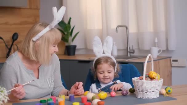 女性と小さな女の子は カウンタートップ上の植物と食器の間に一緒にイースターエッグを飾るキッチンテーブルで瞬間を共有しています — ストック動画