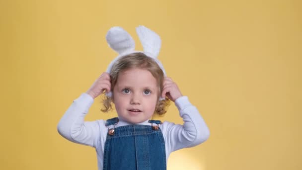 一个在兔子耳朵里快乐地蹒跚学步的小孩 在黄色的背景上微笑着 做着一个笨重的手势 她用耳朵玩耍 享受闲暇时光 像个小宝宝一样玩得很开心 — 图库视频影像