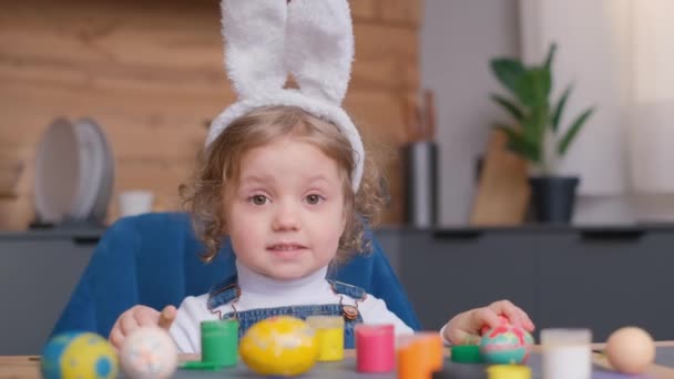 バニーの耳を持つトッドラーは ハウスプラントやフラワーポットに囲まれたイースターエッグを飾るテーブルに幸せに座っています 彼女はカラフルな卵で働くように笑顔を共有しています — ストック動画