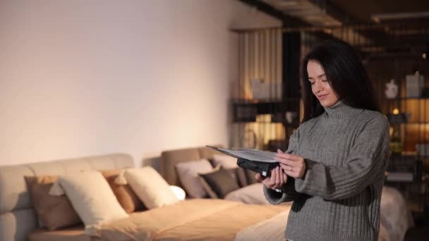 魅力的なブルネットは モダンなインテリアを備えた部屋の背景にある家具生地の紙のカタログを見ます 家のための柔らかい家具の選択と購入 — ストック動画