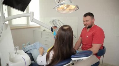 Genç ve çekici bir dişçi dişçi, dişçi koltuğunda otururken güzel bir esmer kadın hastayla konuşuyor. Ağız boşluğundaki hastalıkların tedavisinde modern yaklaşımlar