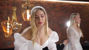 İki gelin bir düğün salonunda gelinlik deniyor. Gösteri, gelinlik galerisi. Mankenler gelinlik gösterisi için hazırlanıyor. Gelin saçını düzeltir.