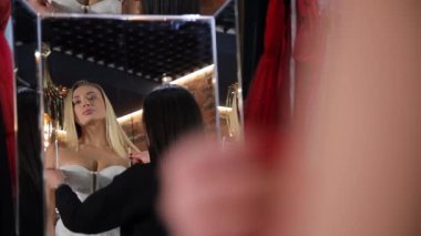 Gelinlik seçen zarif bir gelinin yakın çekimi ve gelinlik seçmesine yardım eden bir satış danışmanı. Gelin aynanın önünde, düğün salonunun soyunma odasında..