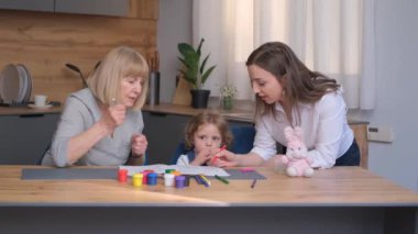 İki kadın ve küçük bir kız, ahşap bir masada boş boş oturuyorlar. Masa takımı, bitkiler ve sanatla çevriliyken gülücüklerini ve sohbetlerini paylaşıyorlar.