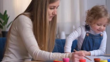 Bir kadın ve küçük bir kız masadaki eğlenceli bir resim etkinliğini paylaşıyorlar, parmakları renkli boyalarla kaplıyken sohbet ediyorlar ve heyecanla el kol hareketi yapıyorlar.