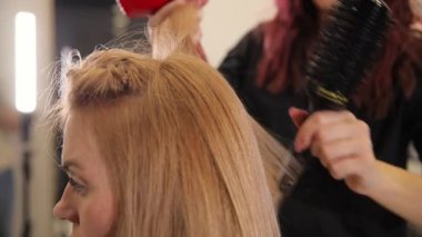 İşteki kuaför kız. Kuaför kız, uzun sarı saçlı, saç kurutma makinesi ve saç fırçasıyla bir kadına şekil veriyor. Profesyonel bir kuaförden saç stili.