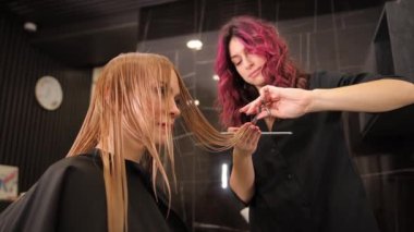Kıvırcık kızıl saçlı kuaför kız sarışın bir müşterinin saçını kesiyor. Güzellik salonunda uzun saçlı profesyonel bir saç kesimi. Saç kısalması. Sağlıklı görünen saçlar.