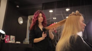 İşteki kuaför kız. Kuaför kız, uzun sarı saçlı, saç kurutma makinesi ve saç fırçasıyla bir kadına şekil veriyor. Profesyonel bir kuaförden saç stili.