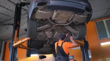 Otomotiv lastiği üzerinde çalışan bir adam bir garajda asansörün altında, otomotiv dışı, tampon, otomotiv aydınlatma ve otomotiv parçaları ile çevrili.