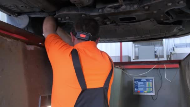 スポーツウェアの男性は 個人的な保護装置のためのヘルメット付きのガレージで車の下で作業しています 彼は自動車のタイヤを修理するときに彼の袖口の腕でジェスチャー — ストック動画