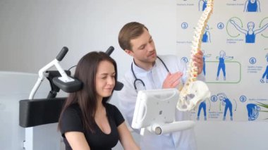 Doktor, makinede oturan bir kadına omurga modelini gösteriyor. Kadın kolu kalçasında dinleniyor ve jesti izliyor.