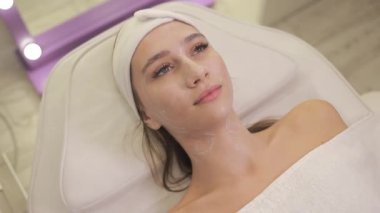 Bir kadın, kafasına bir havlu sarılmış, saçına ve yüzüne odaklanmış bir şekilde spa yatağında yatıyor. Rahatlamış, kaşları ve kirpikleri çok bakımlı.