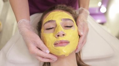 Kadın kaplıcada cildine sarı bir maske taktırıyor. Alnından çenesine kadar yüzünü kapatıyor. Yüzünde bir gülümseme var. Maske nazikçe bir elle uygulanıyor.
