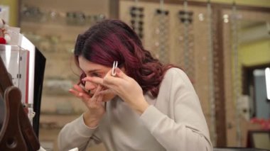 Güzel kıvırcık saçlı genç bir kız bir optik dükkanında aynanın önüne göz lensi takıyor. Görme yetisini geliştirmek için. Göz renginde değişiklik