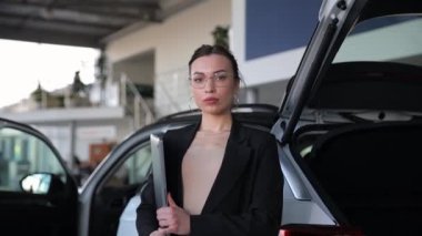 Bir kadın bagajı açık bir aracın yanında, otomotiv tasarımını ve arabanın lastik, ayna ve kapısı gibi dış özelliklerini sergiliyor.