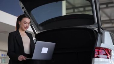 Bir kadın, dizüstü bilgisayarını kullanarak arabanın arkasında oturuyor. Aracın içi, modern otomotiv tasarımı ve aydınlatma ile aydınlatılır.