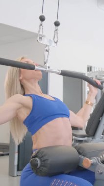 Bir kadın spor salonunda kaslarını çalıştırmak için bir makine kullanıyor. Uygun bir duruşla oturur, karnını, göğsünü, kalçalarını ve kollarını çeşitli egzersizlerle meşgul eder.