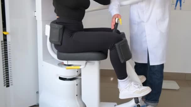 一位女士坐在椅子上 椅子看起来像运动器械 身后站着一位医生 这台机器集中在她的腿 大腿和膝关节上 — 图库视频影像