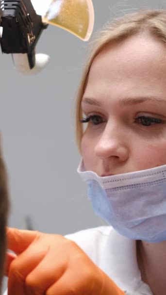 女性歯科医は 患者の歯を調べながら保護マスクとオレンジの手袋を着用しています 彼女の髪は慎重に口 および周囲の顔の特徴を検査するので 背中に縛られています — ストック動画