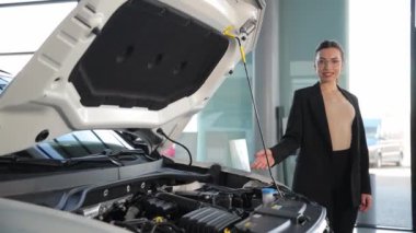 Bir kadın kaputu açık bir şekilde bir motorlu aracın önünde duruyor, otomotiv tasarımı ve ışıklandırması sergiliyor. Arabaya doğru bir jest yapıyor, dış görünüşünü vurguluyor.