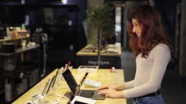 İnce, güzel bir kıvırcık kız dijital ekipman mağazasında dizüstü bilgisayarda çalışıyor. Cep telefonu sergisinin arka planındaki kız satıcı.