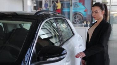 Bir kadın galeride lüks bir arabanın yanında duruyor, otomotiv tasarımına, dış görünüşüne, aydınlatmasına, lastiklerine, kapılarına ve aynalarına hayran.