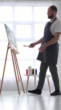 Bir adam bir bacağının üzerinde duruyor, dizini hafifçe büküyor ve diğer bacağının üzerinde dengede duruyor, bir sehpada resim yaparken.