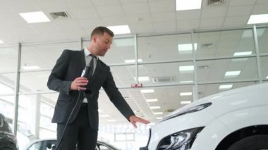 Modern bir araba galerisinde arabanın sunumu. Genç ve çekici bir danışman elektrikli bir arabayı nasıl şarj edeceğini gösterir. Arka planda güzel bir araba satıcısı.
