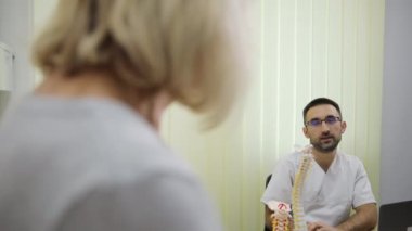 Resmi kıyafetli bir doktor bir hastasıyla masada oturup sohbet ediyor. Doktorun jesti cana yakın, sakalı düzgün taranmış, tıbbi olaya biraz sıcaklık ve eğlence katmış.