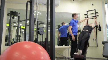 Bir erkek, diz egzersizi yapan bir kadına spor aletlerini kullanarak kalçalarını güçlendirmesi ve fiziksel kondisyonunu iyileştirmesi için yardımcı oluyor.