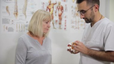Resmi giyinmiş bir adam, bir sanat etkinliğinde bir kadınla konuşurken bir insan kafatası modeliyle el kol hareketi yapıyor. Sohbet arka planda görsel sanatlar ve gözlüklerle eğlenceli ve ilgi çekici görünüyor.