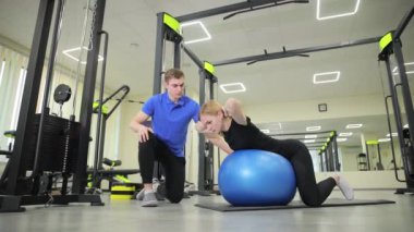 Bir adam pilates topuyla bir kadına yardım ediyor, bacağına ve bacak kaslarına odaklanıyor, spor kıyafetlerini spor salonunda giyip spor salonunda spor yapıyor.