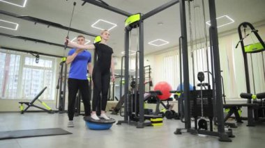 Bir erkek ve kadın spor salonunda birlikte çalışıp, ortak egzersizlere odaklanıyorlar. Daha sonra, kadın ağırlık eğitimi için çeşitli egzersiz makineleri ve ekipmanlar kullanan bir fitness eğitmeni ile antrenman yapar.