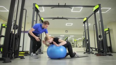 Bir adam spor salonunda mavi egzersiz topu üzerinde egzersiz yapan bir kadına yardım ediyor. Spor malzemeleriyle yerde eklemler, kollar, bacaklar, bacaklar ve dizler üzerinde çalışıyorlar.