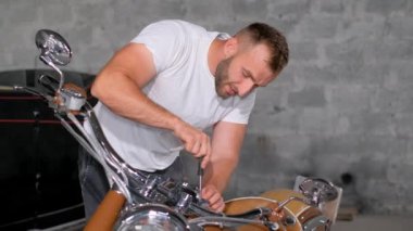 Otomobil parçaları ve aletlerle çevrili bir adam garajda motosikletini tamir ediyor. Arka planda çalan müziğin sesi çevreye eklenir