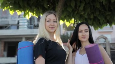 İki güzel, atletik kadın bir yaz günü bir ağacın altında jimnastik paspasıyla kameraya bakarak poz veriyor. Eğitimden önce spor aletleri olan ince bir kadın.