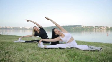İki genç kadın göl kıyısında gökyüzünün arka planında otururken yoga yapıyorlar. Yoga, sakin bir gölün kıyısında, temiz havada. Sağlık kavramı.