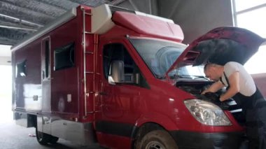 Bir tamirci, taşımacılık için kullanılan kırmızı bir karavanın motorunu tamir ediyor. Adam araçlara ve motorlara odaklanmış durumda.