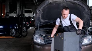 Bir adam garajda kapüşonlu bir aracı tamir ediyor. Arabanın motoru ve dış kısmı üzerinde çalışıyor. Otomotiv aletleri ve ekipmanlarla çevrili.